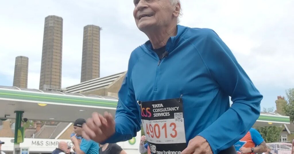 Pessoa correndo com número de inscrição em uma maratona, fundo urbano com prédios e posto de combustível.