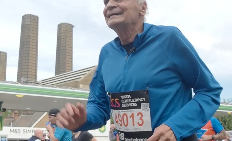 Pessoa correndo com número de inscrição em uma maratona, fundo urbano com prédios e posto de combustível.