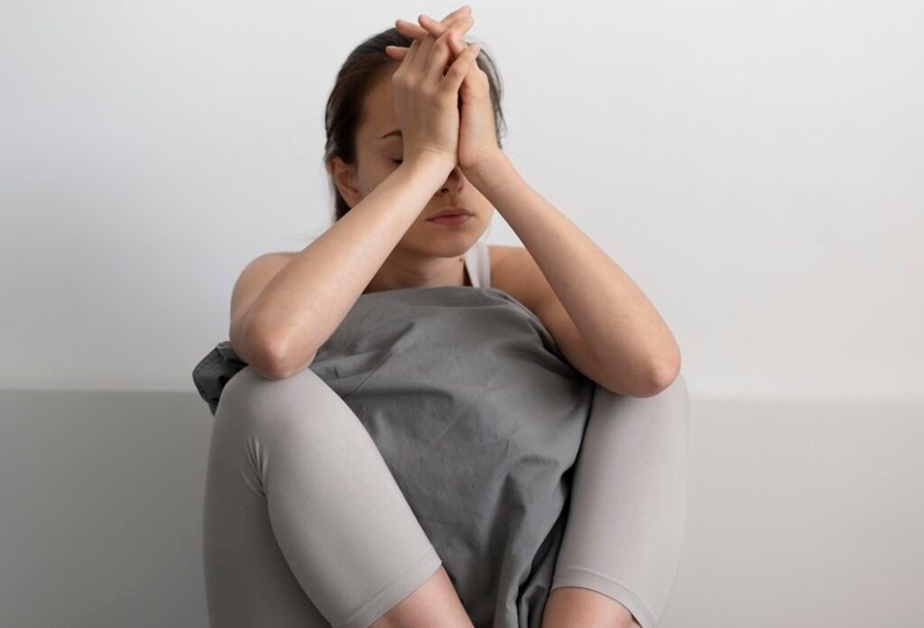 Pessoa usando roupas casuais sentada com os joelhos dobrados, fundo claro.