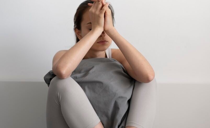 Pessoa usando roupas casuais sentada com os joelhos dobrados, fundo claro.