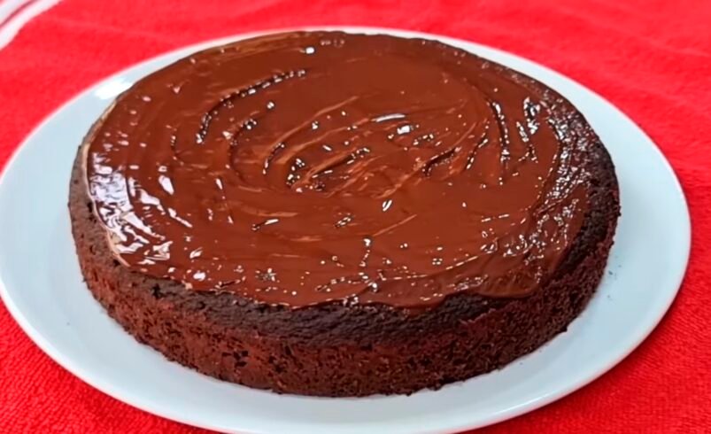 Bolo de chocolate com cobertura brilhante sobre prato branco e fundo vermelho.
