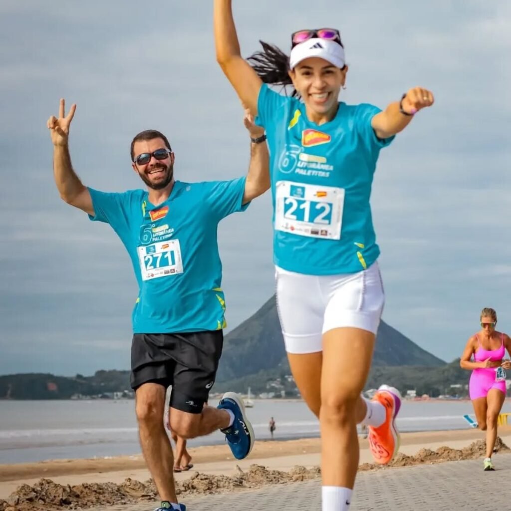 Duas pessoas correndo em uma praia com montanhas ao fundo, dia ensolarado.