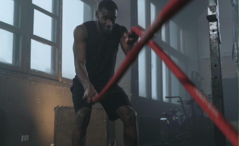 Homem em ambiente de academia treinando com corda naval vermelha.