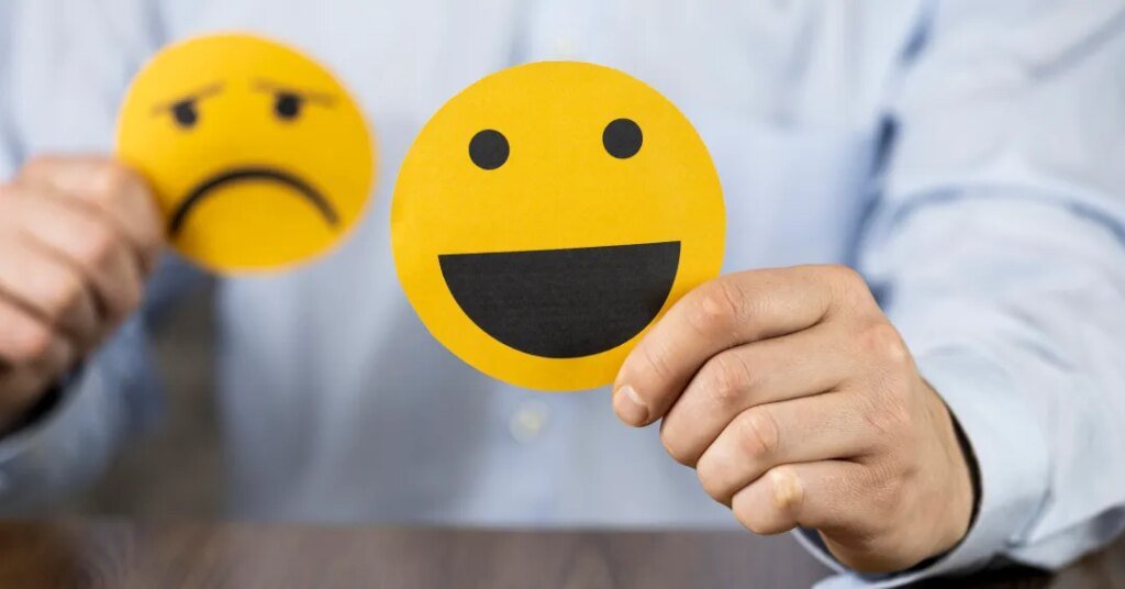 Pessoa segurando emojis de rosto feliz e triste em fundo neutro.
