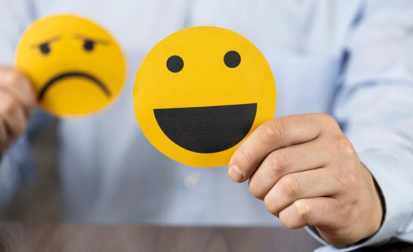 Pessoa segurando emojis de rosto feliz e triste em fundo neutro.