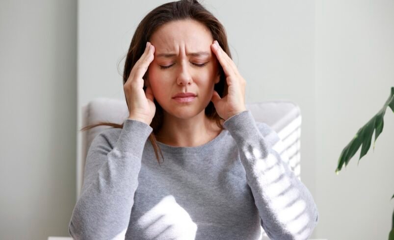 Pessoa em ambiente claro segurando a cabeça com as mãos, vestindo blusa cinza.