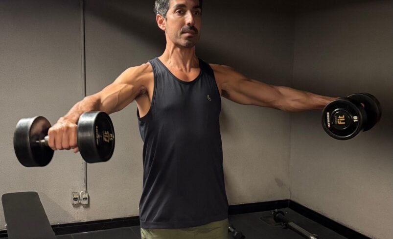 Pessoa realizando elevação lateral com halteres em uma academia.