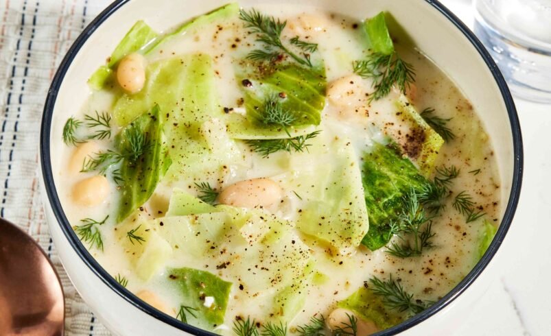 Tigela com sopa de repolho, feijão branco e ervas frescas ao lado de uma colher.