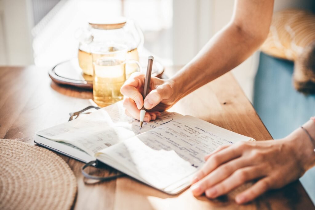 Mão escrevendo em caderno aberto sobre mesa com chá ao fundo.