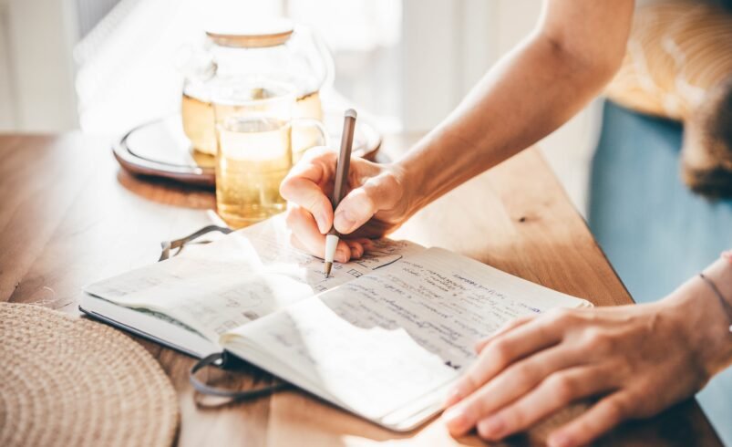 Mão escrevendo em caderno aberto sobre mesa com chá ao fundo.
