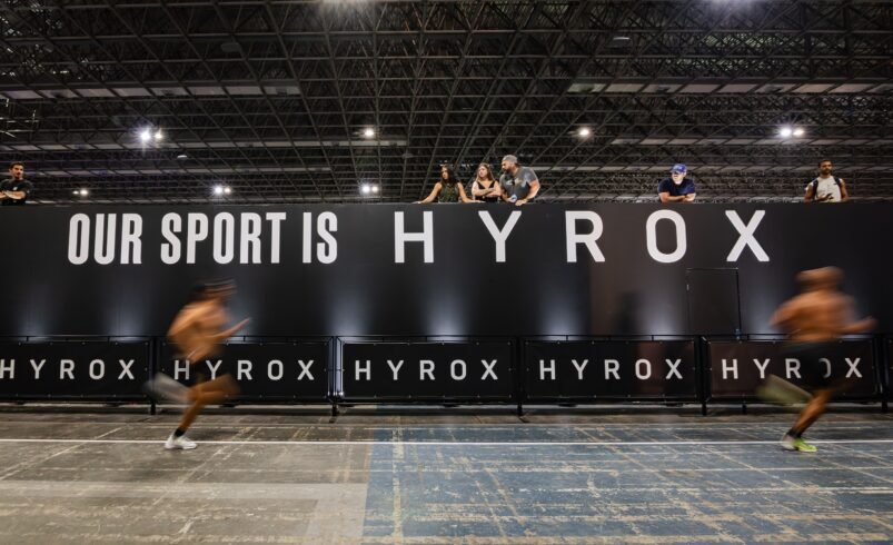 Corredores em pista interna de evento Hyrox com banner destacado.
