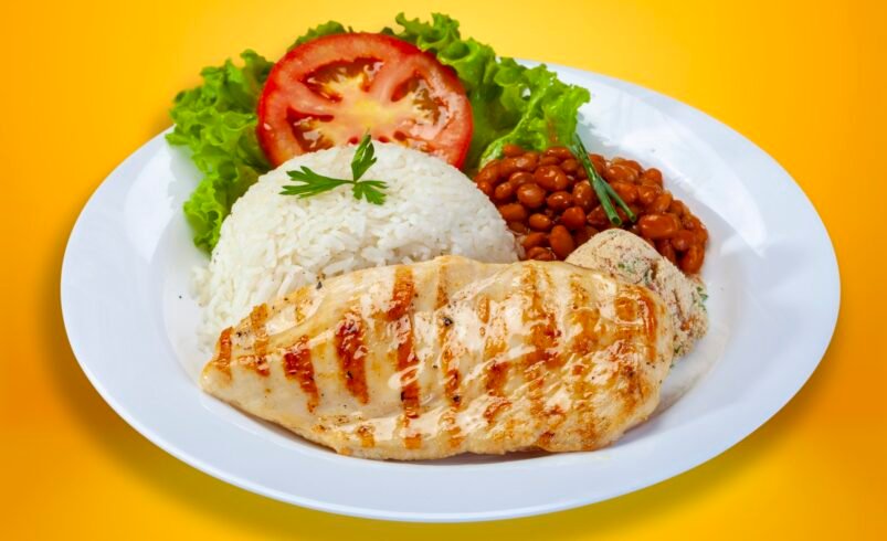 Prato de frango grelhado com arroz branco, feijão e salada sobre fundo amarelo.