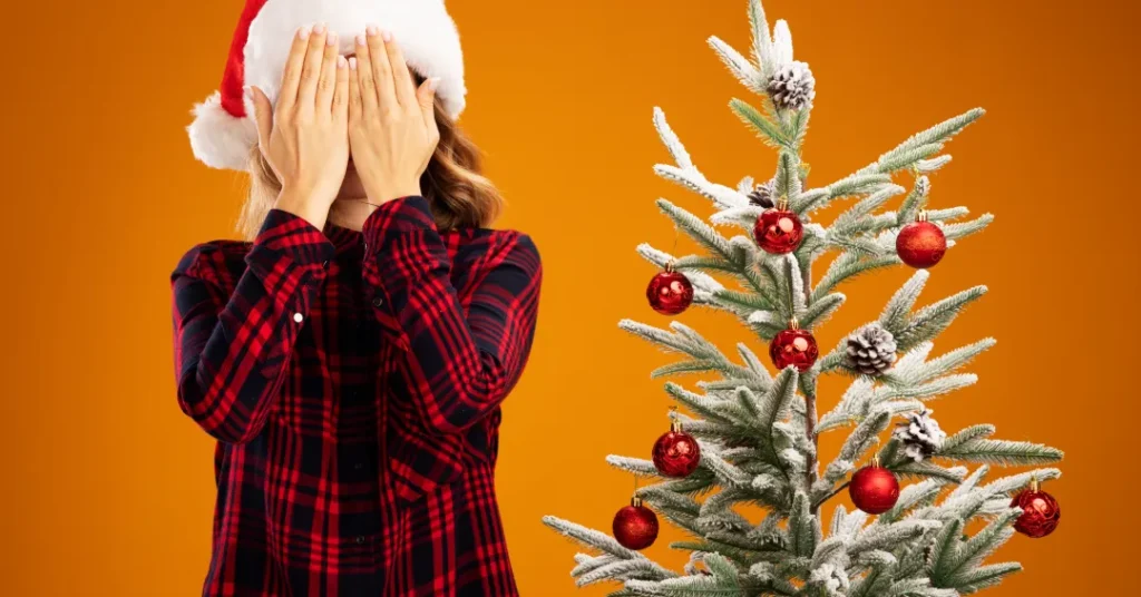 Mulher com gorro de Papai Noel cobrindo o rosto ao lado de árvore de Natal decorada.