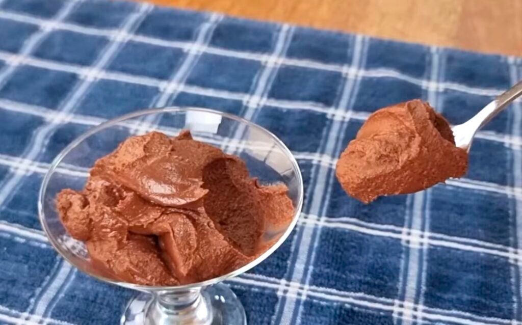 Só 2 ingredientes! Delícia sem açúcar, fácil e rápida - Sem leite condensado ou creme de leite 1 Taça de mousse de chocolate sobre toalha azul, com colher servindo.