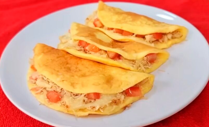 Três crepiocas recheadas com frango desfiado e tomate em prato branco sobre toalha vermelha.