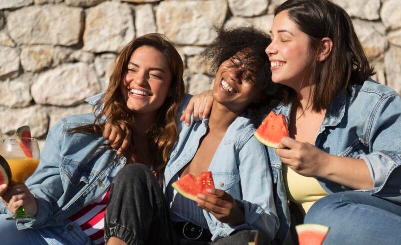 Três pessoas juntas ao ar livre segurando fatias de melancia, com um muro de pedra ao fundo.