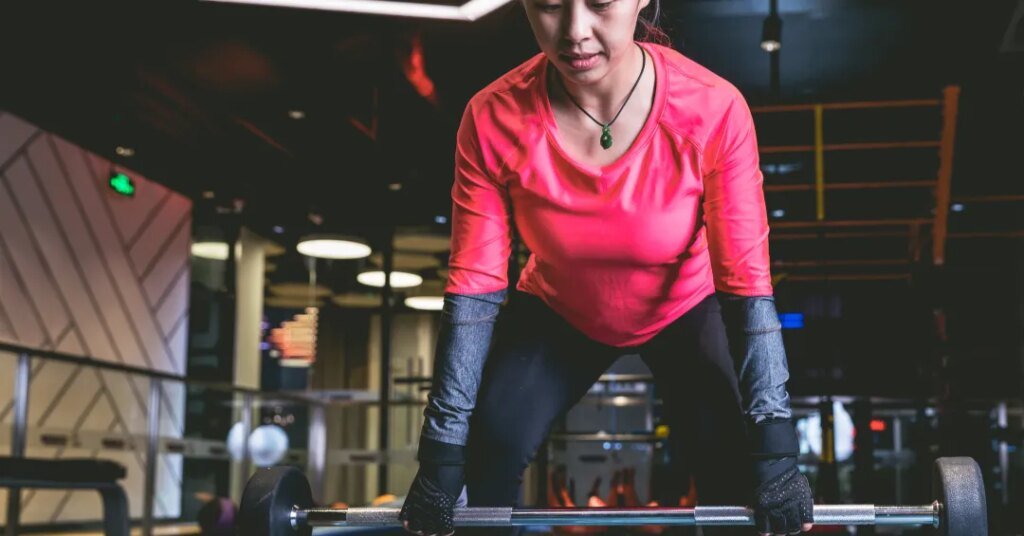 Pessoa usando roupa esportiva, levantando barra em academia iluminada.