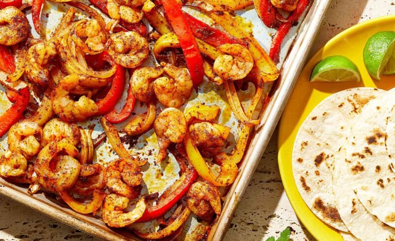 Fajitas de camarão douradas com pimentões coloridos em uma assadeira e tortilhas ao lado.