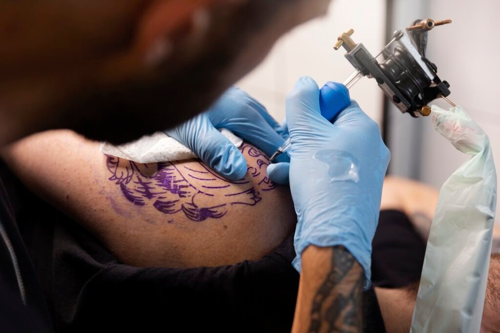 Tinta de tatuagem pode afetar defesa do corpo e resposta a vacinas, afirma estudo 1 Tatuador com luvas azuis criando uma tatuagem roxa em um braço.