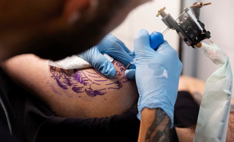 Tatuador com luvas azuis criando uma tatuagem roxa em um braço.