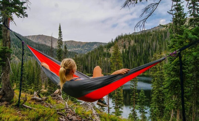 Pessoa relaxando em uma rede vermelha entre árvores em área montanhosa.