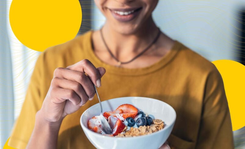 Pessoa sorrindo segura tigela de frutas, granola e iogurte.