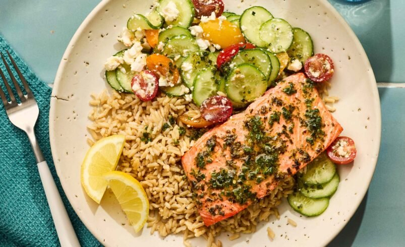 Prato com salmão grelhado, arroz integral, salada de pepino e tomate cereja acompanhado por fatias de limão.