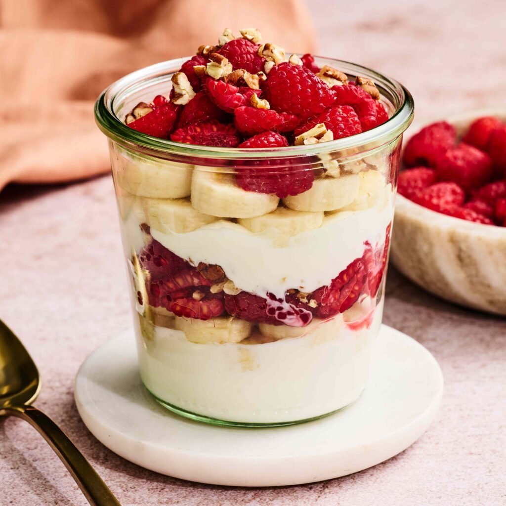 Mais de 15 receitas de café da manhã adequadas para diabetes que não são aveia 1 Copo com iogurte, banana, framboesa e granola decorado sobre suporte branco.