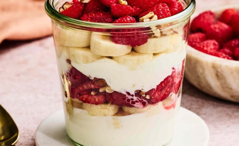 Copo com iogurte, banana, framboesa e granola decorado sobre suporte branco.