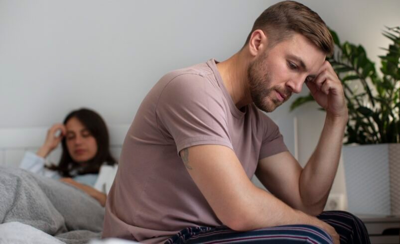 Homem com expressão preocupada sentado em uma cama, ao lado de plantas.
